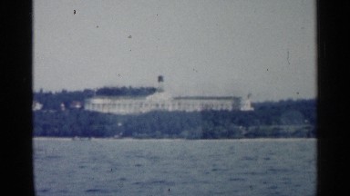 1960: view from a boat of a big, white mansion surrounded by trees on the shore