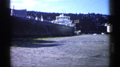 1961: long and flat-bottom ship sits on grass in front of mountainside town