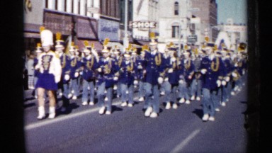 1961: band in blue marches in city parade with bent arms holding instruments