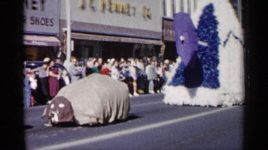 1961: old firetruck being driven by firefighters in a parade with other floats