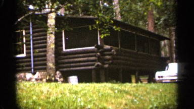 1965: house made with wooden logs,surrounded with trees, a car parked on green