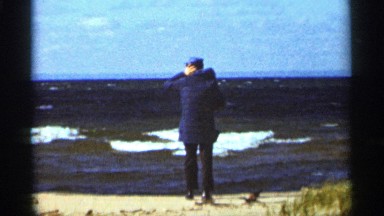 1965: man in an overcoat and brimmed cap, standing on a windy day by the ocean