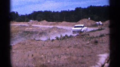 1965: silver automobile cruises up a dirt road away from the camera