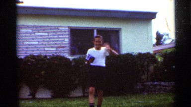 1963: Boy Waves and Takes a Picture in Backyard While Man in Hat Pushes Stroller