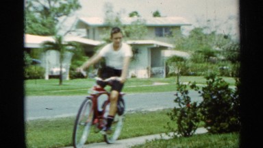 1963: boy riding his red bicycle around the street towards his house