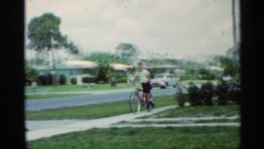 1963: young boy waves to the camera as he is standing up riding a ten speed bike
