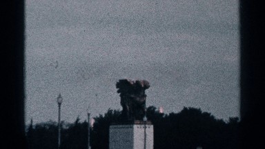 1964: a large bronze statue sits atop a pedestal in a city as cars drive past