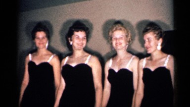 1958: four women wearing black dresses sing together holding an object