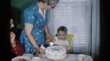 1958: Grandmother Seating Birthday Boy in Front of His Cake While Family Celebrates