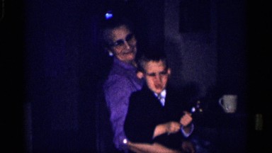 1959: an older lady in glasses holds a boy as he plays and waves to the camera