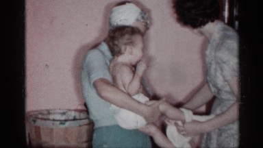 1955: infant being dressed up by her nanny, while a lady smiles at the baby.