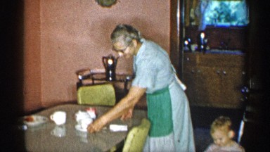 1955: Interior Home Scene with Woman in Apron, Child Playing, and Adults Setting Table