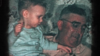 1955: man with glasses holding a child in a blue shirt reaching for a dog
