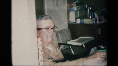 1959: interior, showing bespectacled person sitting next to an empty chair.