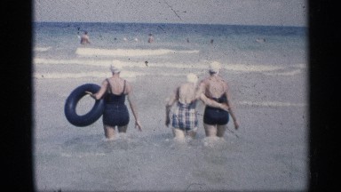 1959: three woman wearing one piece bathing suits wade out into the water