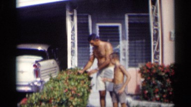 1959: a man and his son stand outside of their house shirtless picking a flower