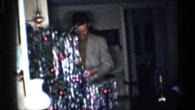 1959: man decorating a christmas tree that has presents underneath it.