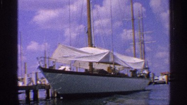 1959: a sailing ship with a very tall mast docked at a port in calm waters