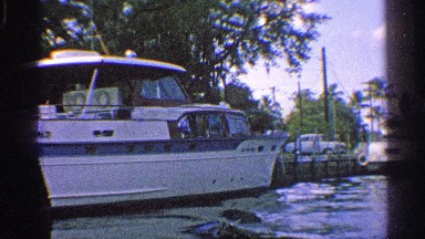 1959: a large cruiser is tied to a dock dotted with trees and with vehicle