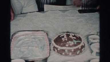 1959: cutting a birthday cake on a white table while being surrounded by friends
