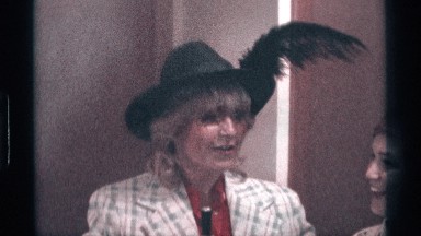 1976: woman smiling while standing next to a woman wearing a fancy black hat