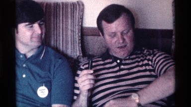 1976: older man with sideburns and striped polo talking into mic with younger