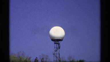 1969: Water Tower is a Supported White Sphere Above Trees and Against Sky