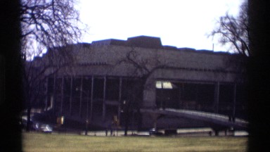 1966: building at the main and ground car moving men walking at the place trees