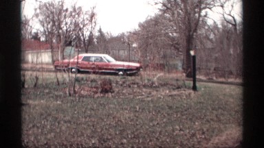 1969: red car rounds a curve on country road and lit headlights are revealed