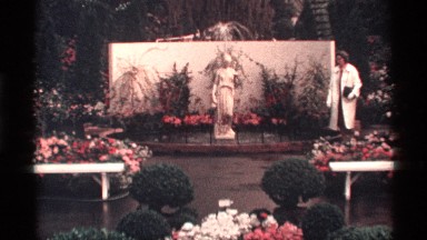 1968: an older lady walking through a beautiful garden with flowers, shrubs and