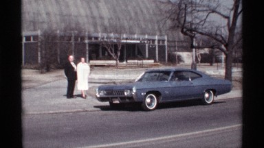 1968: man opens passenger door for woman after they pose by steel-blue car