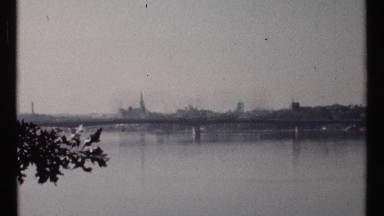 1967: long bridge crosses river that has city buildings as a distant border