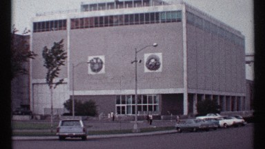 1967: vehicles parked and moving on the road in front of a large spectacular