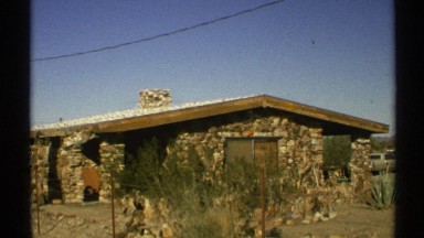 1972: a rock house sits behind a fence and scrub brush in a desert environment