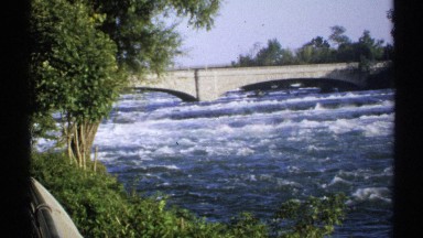 1971: bridge side area with trees, water flowing over a big river a distant