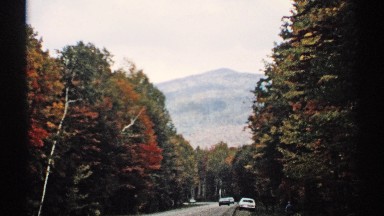 1971: street lined with trees, with parked & passing cars and hills in