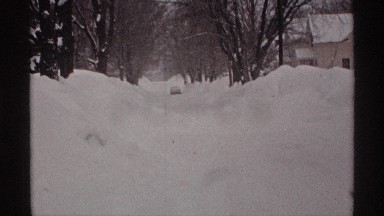 1971: snow filled road with trees on each side and a car parked far away at the