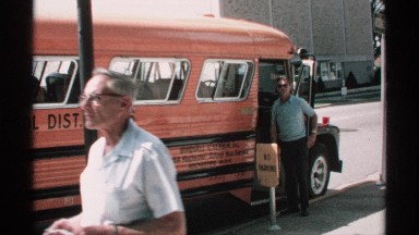 1970: man paces by open door of orange bus while older pedestrian passes by