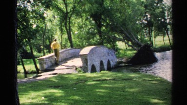 1970: Man Walking Near Stone Bridge, River, and Trees in Minnesota