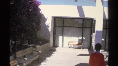1975: Woman Wearing Orange Jacket Walks by Tiered Fountain in Building Plaza