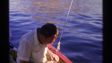 1975: two men baiting a fishing line in a small boat with a large rock structure
