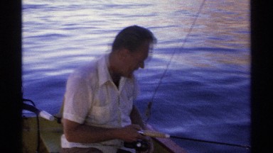 1975: man on small boat holds and looks down at fishing rod cast into water