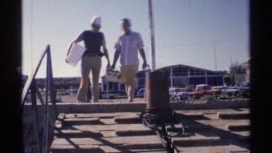 1975: man lifts bag from cardboard box while other man holds insulated bin