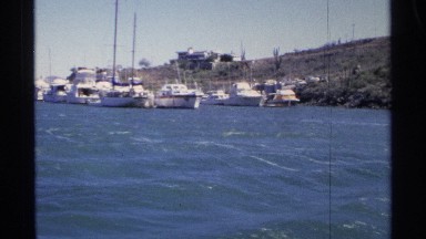 1975: wide and short building is on sloped coast above aligned white boats