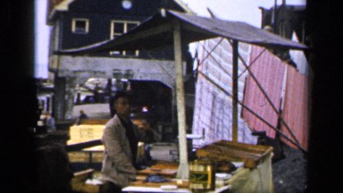 1957: a man is sitting at an outdoor booth that is at the edge of the water