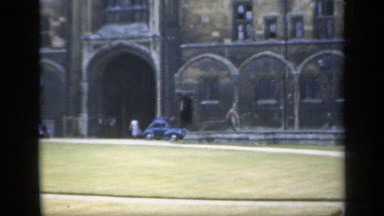 1957: a person is walking outside in front of a tall, old, stone building.