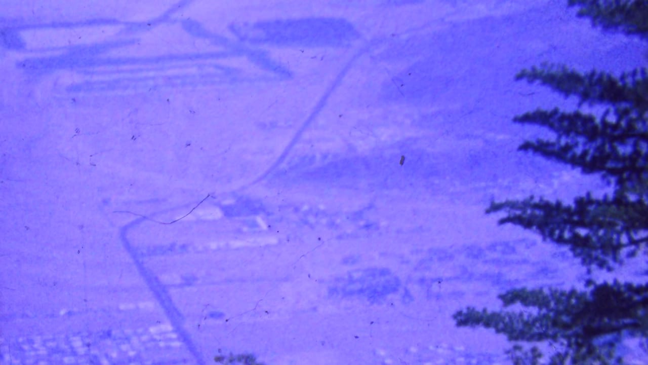 1964 Palm Springs-area 8mm frame showing a hazy elevated view over desert roads and building clusters, partly framed by dark tree branches.