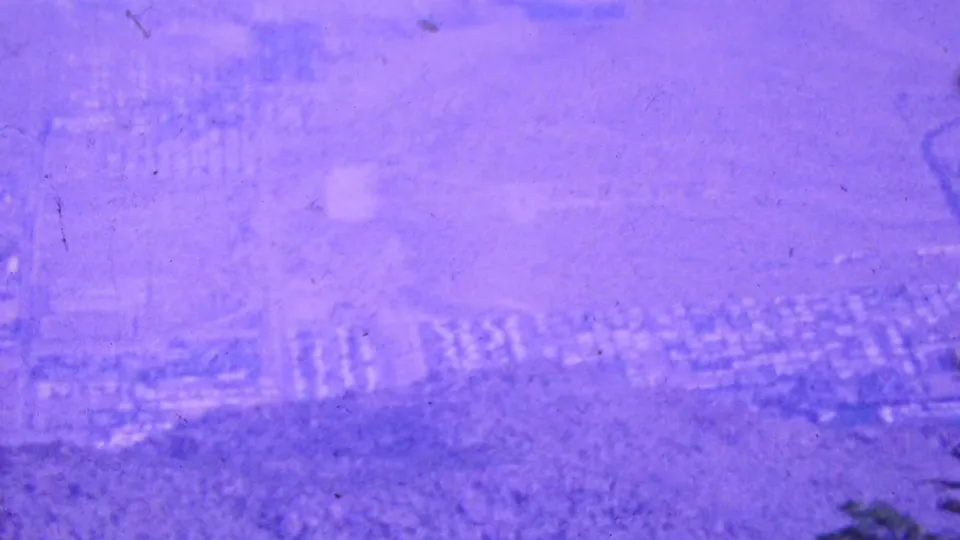 1964 Palm Springs-area elevated view over a hazy desert settlement pattern, with faint roads and building clusters softened by aged 8mm film color shift.