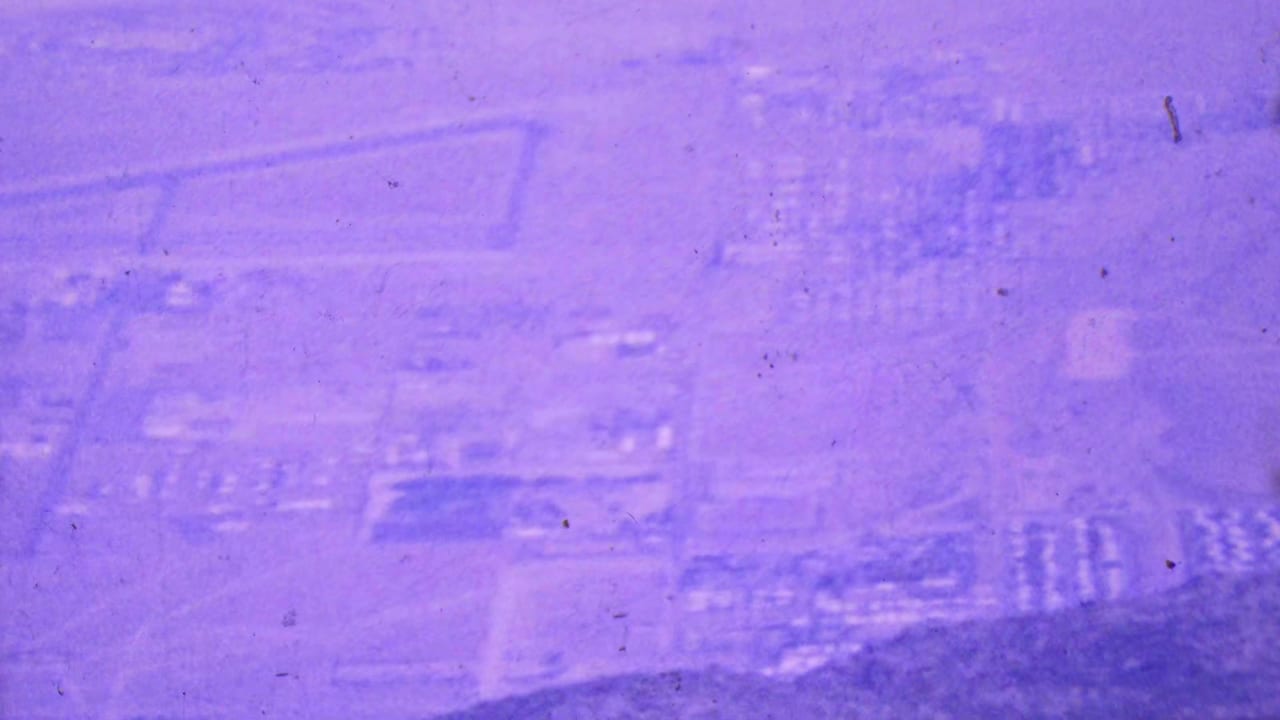 Soft 1964 aerial-tramway view over Palm Springs with faint rectangular land divisions, roads, clustered buildings, and visible film grain and dust.