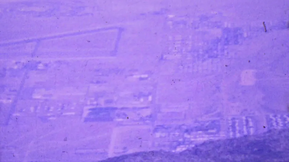 Soft 1964 aerial-tramway view over Palm Springs with faint rectangular land divisions, roads, clustered buildings, and visible film grain and dust.